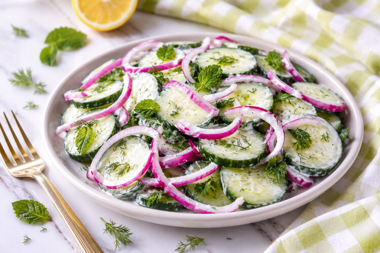 Salade crémeuse de concombres tranchés garnie de fines rondelles d’oignon rouge et de feuilles de menthe fraîche, servie dans une assiette blanche. La salade est enrobée d’une vinaigrette au yogourt et aux herbes, avec un citron et un linge à carreaux vert pâle en arrière-plan.