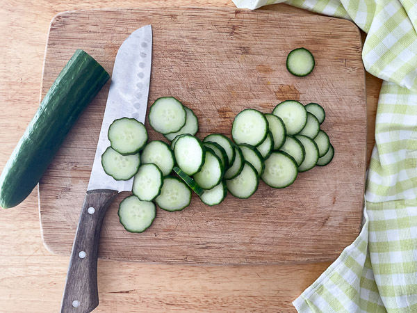 Concombre frais coupé en fines rondelles sur une planche à découper en bois, avec un couteau de cuisine à proximité.