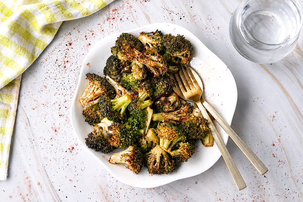 Assiette de brocoli rôti au Air Fryer assaisonné de paprika, d'ail et de gingembre, photographiée en vue de dessus sur une table en marbre blanc avec une fourchette dorée et un verre d'eau.