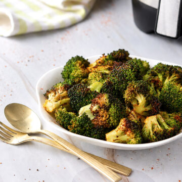 Brocoli rôti au Air Fryer servi dans une assiette blanche, avec des fleurons tendres aux extrémités bien dorées, accompagné d'ustensiles dorés.