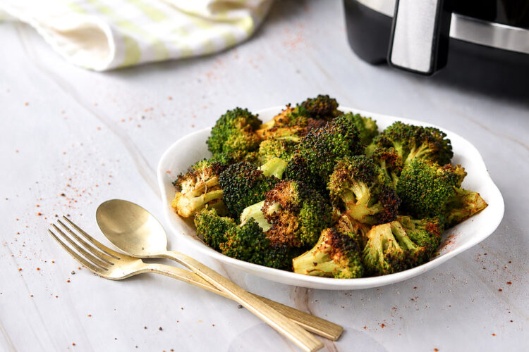 Brocoli rôti au Air Fryer servi dans une assiette blanche, avec des fleurons tendres aux extrémités bien dorées, accompagné d'ustensiles dorés.