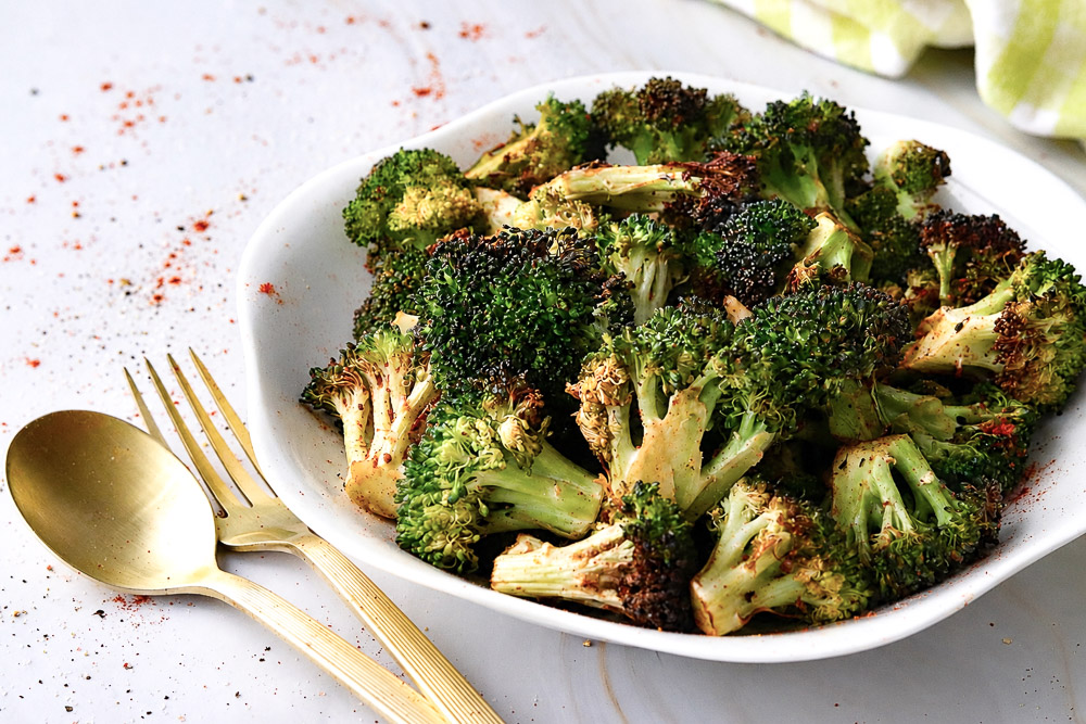 Brocoli rôti au Air Fryer assaisonné de paprika, d'ail et de gingembre, servi dans une assiette blanche sur une table en marbre avec des ustensiles dorés.