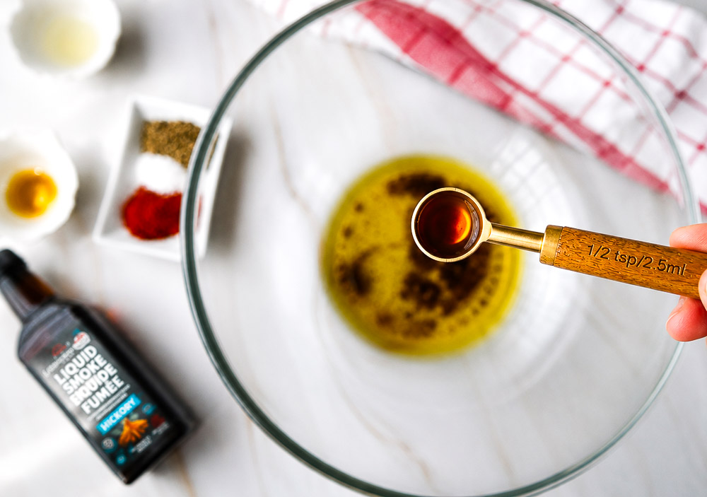 Demi-cuillère à thé de fumée liquide tenue au-dessus d'un bol d'huile d'olive, avec les épices et la bouteille de fumée liquide en arrière-plan.