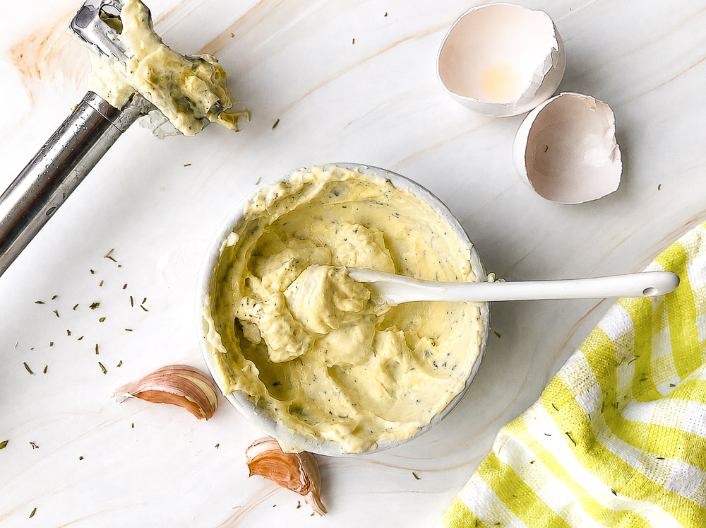 Mayonnaise maison à l'huile d'olive et au thym dans un bol en verre, garnie d'ail frais, avec un mélangeur à immersion recouvert de mayonnaise en arrière-plan.