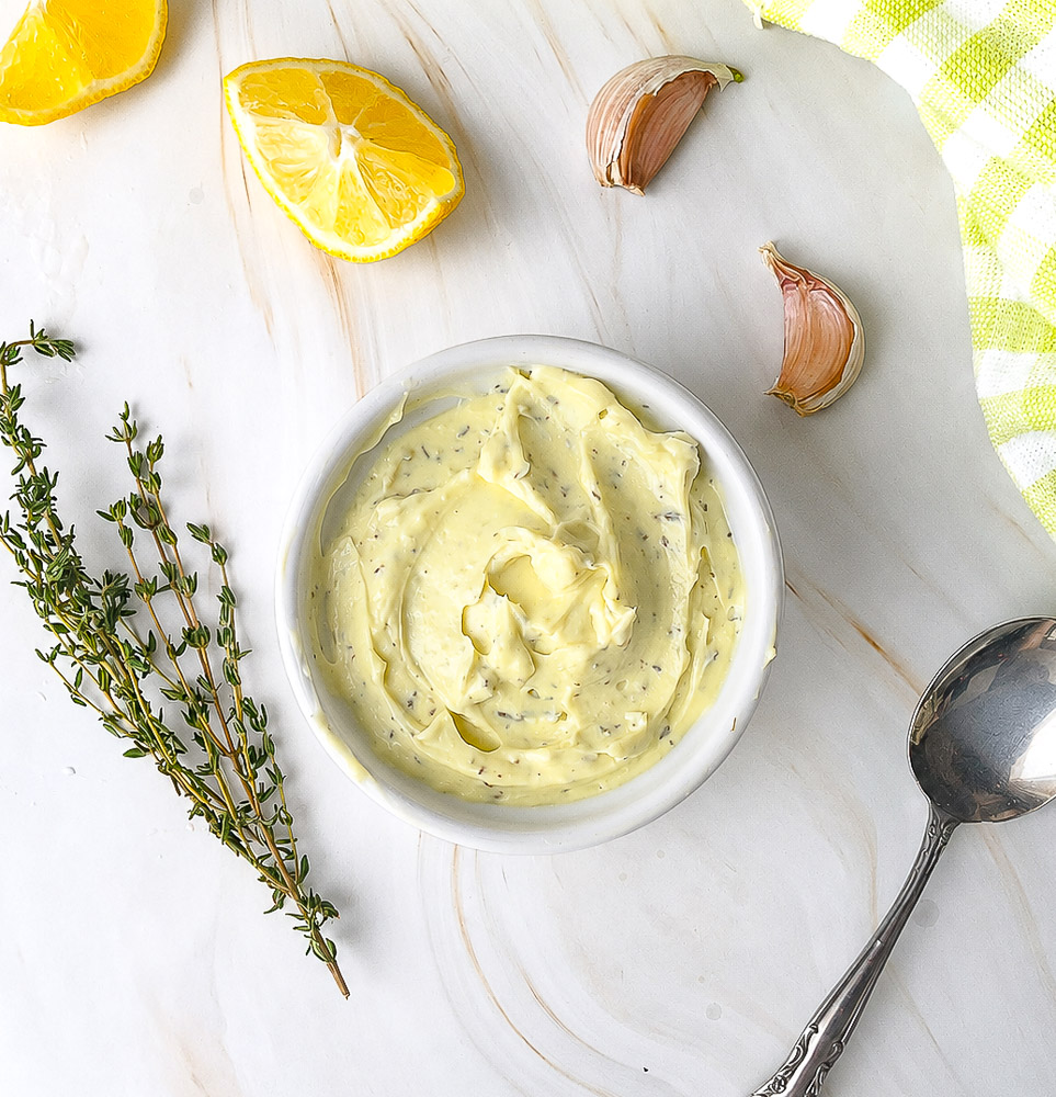 Vue de dessus d'un bol de mayonnaise maison à l'huile d'olive et au thym, accompagné de citron frais, de gousses d'ail et de branches de thym sur un plan de travail clair.