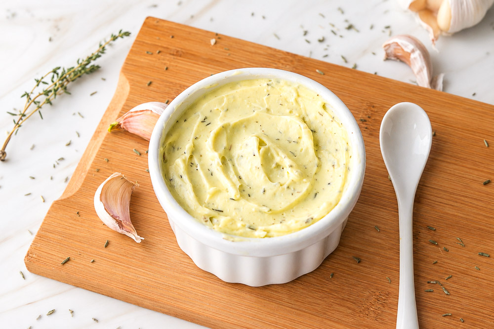Bol de mayonnaise maison à l'huile d'olive et au thym garni de gousses d'ail, présenté dans un ramequin blanc sur une planche de bois avec une cuillère blanche à côté.