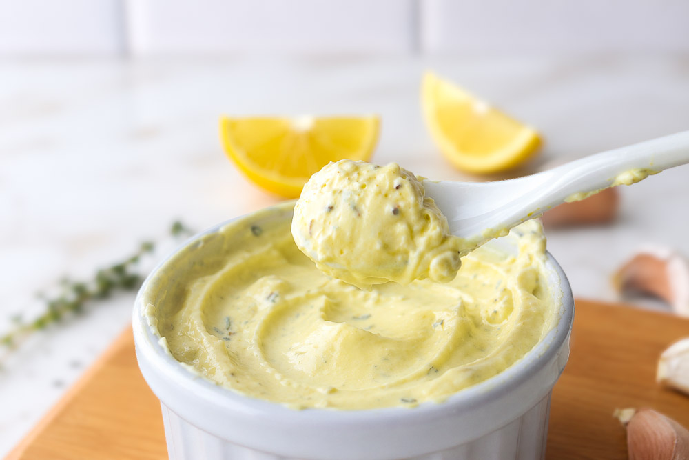 Cuillère blanche remplie de mayonnaise maison à l'huile d'olive et au thym au-dessus d'un ramequin blanc, avec du citron et des gousses d'ail en arrière-plan.