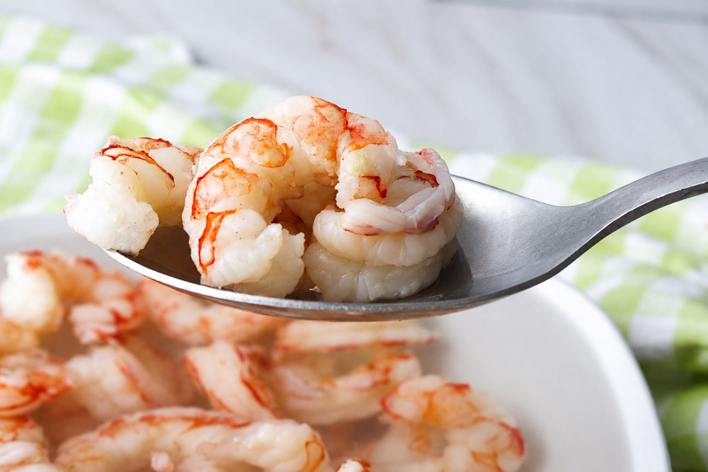 Gros plan de crevettes pochées tendres et juteuses déposées dans une cuillère, avec un bol de crevettes en arrière-plan sur une surface en marbre clair.