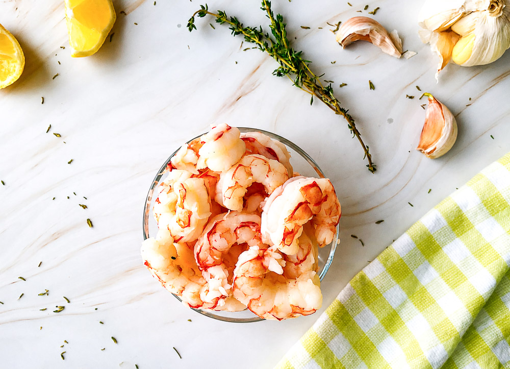 Bol rempli de crevettes pochées vues de haut, accompagné de citron, d'ail et de thym frais sur une surface en marbre clair.