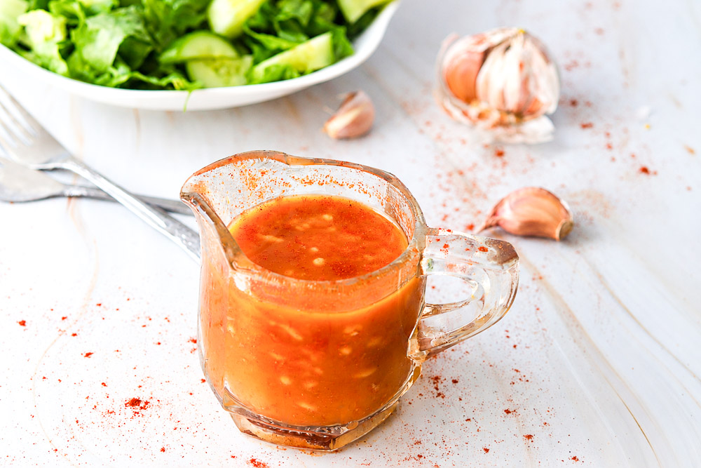 Vinaigrette maison au paprika fumé et à l’ail servie dans un petit pichet en verre, avec salade verte fraîche, gousses d’ail et paprika fumé en arrière-plan.