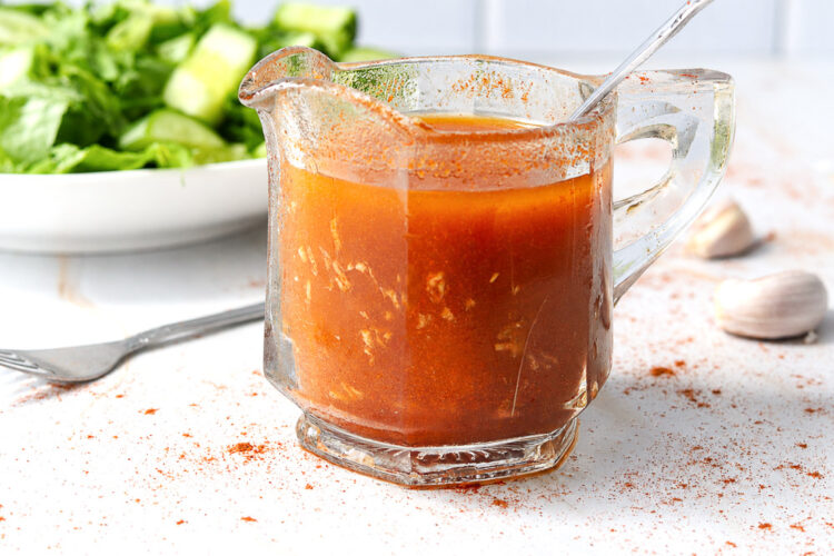 Vinaigrette maison au paprika fumé et à l’ail servie dans un petit pichet en verre, avec salade verte fraîche et gousses d’ail en arrière-plan.