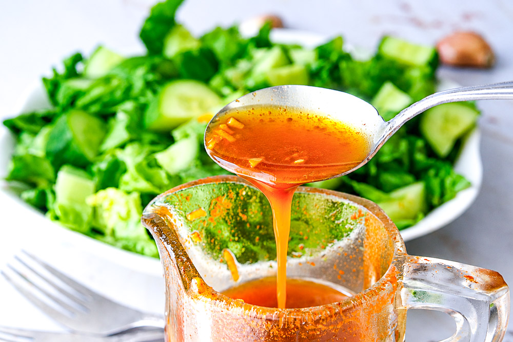 Cuillère de vinaigrette au paprika fumé et à l’ail au-dessus d’un pichet en verre, avec salade verte fraîche en arrière-plan.