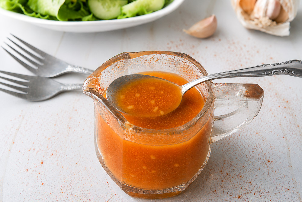 Vinaigrette maison au paprika fumé et à l’ail servie dans un petit pichet en verre, avec cuillère, salade verte fraîche et gousses d’ail en arrière-plan.